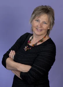 Frau mit kurzen blonden Haaren, Arme verschränkt, schwarzes Oberteil, lächelt.